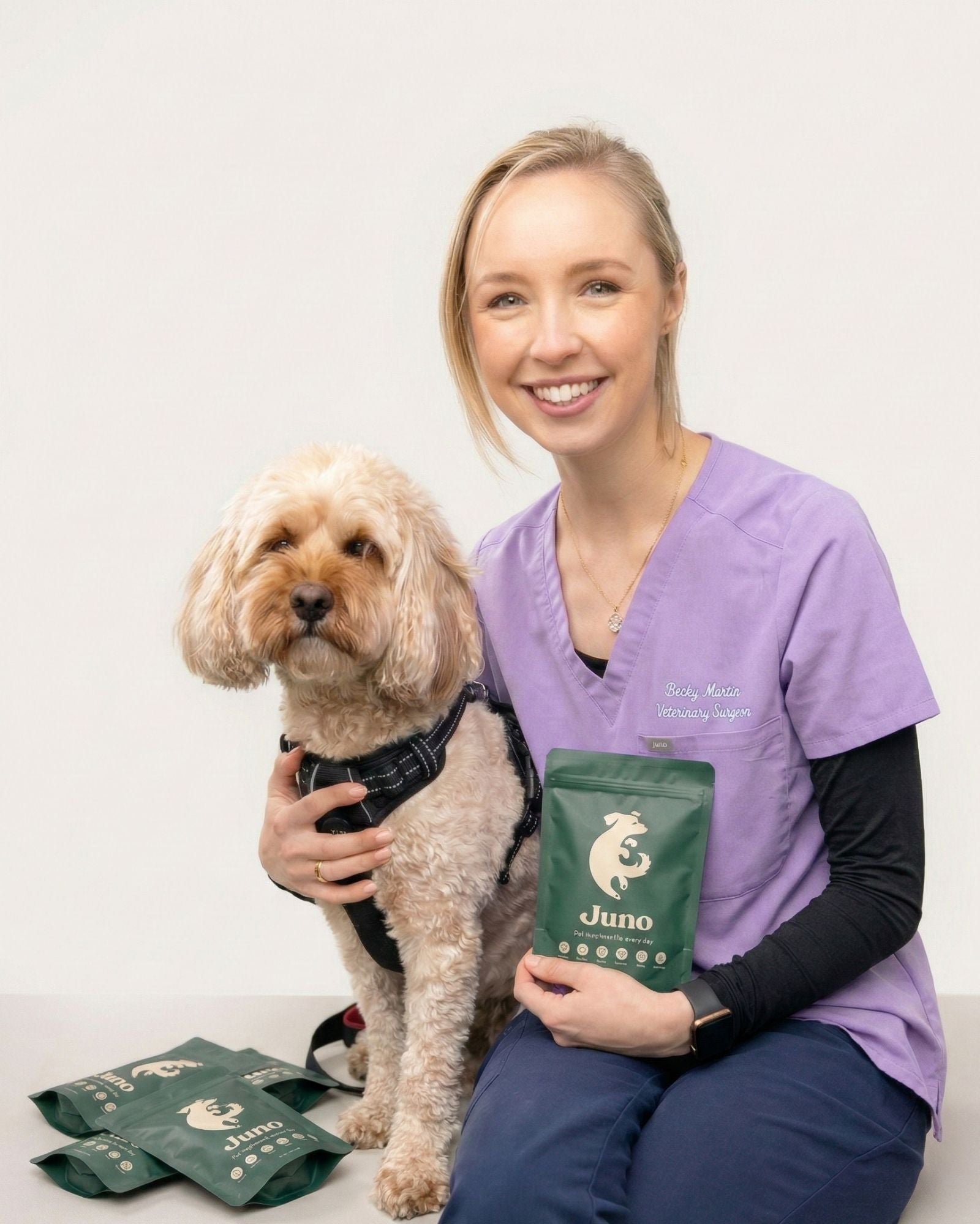 Dr Rebecca Massie in purple veterinary scrubs holding a dog and a green bag with 'Juno' branding on a white background.